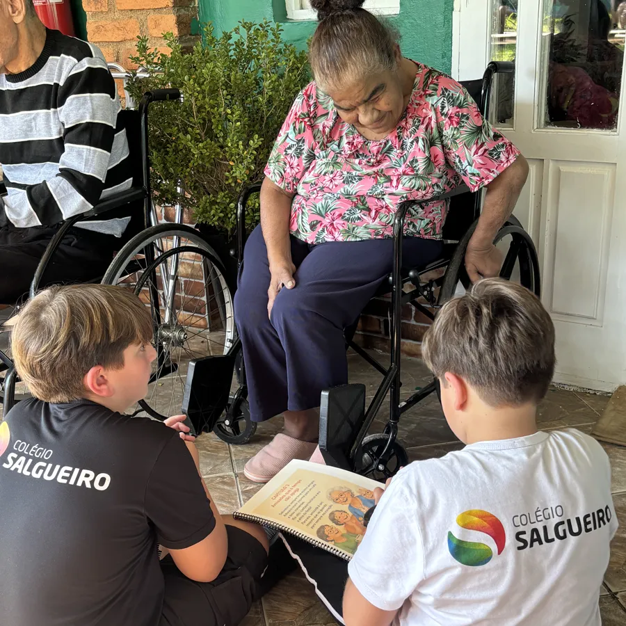 Alunos do 5º ano sentados no chão lendo um livro para uma moradora do lar de idosos