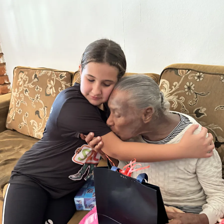 Aluna entregando uma sacola de presente e abraçando uma idosa no sofá