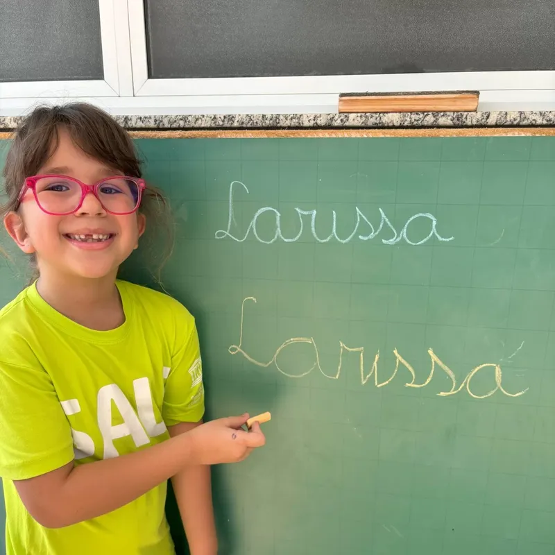 Aluna do 1º ano sorrindo ao lado do próprio nome escrito em letra cursiva na lousa