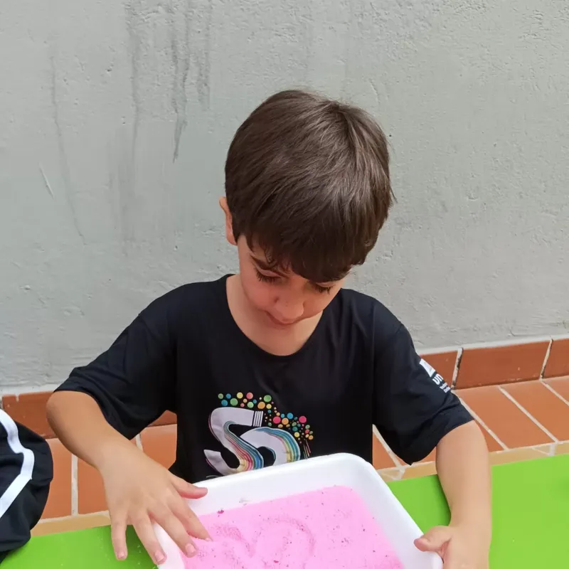 Aluno do 1º ano treinando letras com o dedo em bandeja de areia colorida
